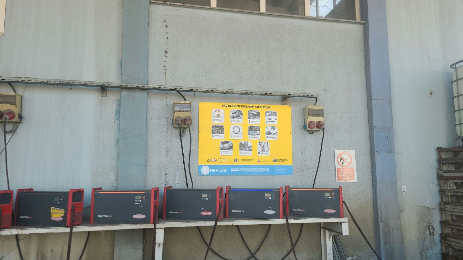 Duvar önündeki tezgâhta yan yana dizilmiş forklift akü şarj redresörleri ve akü bakım talimatı panosu.