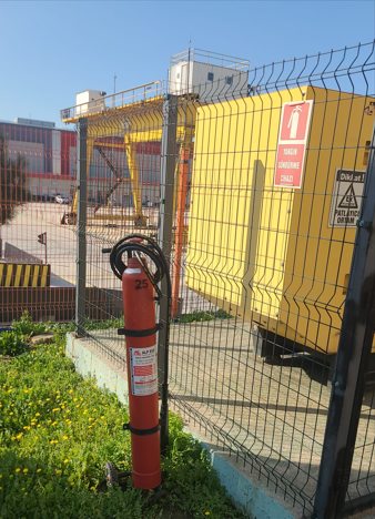 Tesis dışındaki sarı konteyner tipi jeneratör kabini; önünde yangın söndürme tüpü ve uyarı levhaları.
