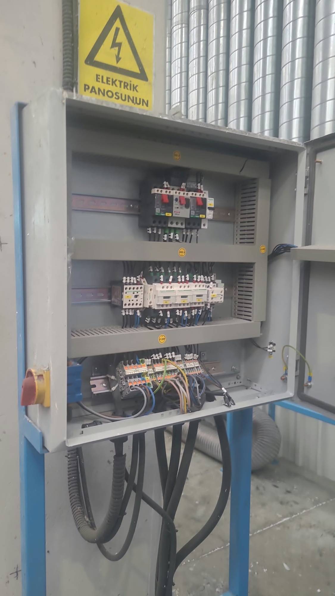 Sahada bir elektrik panosu; devre kesiciler, kontaktörler ve terminal blokları düzenli şekilde monte edilmiş, üzerinde 'Elektrik Panosunun' uyarı etiketi.